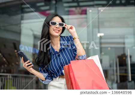 Asian woman wearing sunglasses holding phone carrying shopping bags standing in front of mall plaza. Asian woman wearing sunglasses holding phone carrying shopping bags standing in front of mall plaza. 136648596