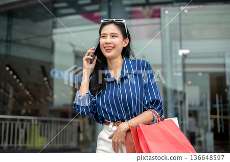 Asian woman holding or talking on phone while carrying shopping bags standing in front of mall plaza Asian woman holding or talking on phone while carrying shopping bags standing in front of mall plaza 136648597