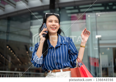 Asian woman talking on phone call and carrying shopping bags while standing in front of a mall plaza Asian woman talking on phone call and carrying shopping bags while standing in front of a mall plaza 136648598