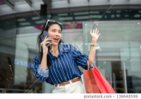 Asian woman talking on phone and carrying shopping bags while standing in front of mall's glass wall Asian woman talking on phone and carrying shopping bags while standing in front of mall's glass wall 136648599