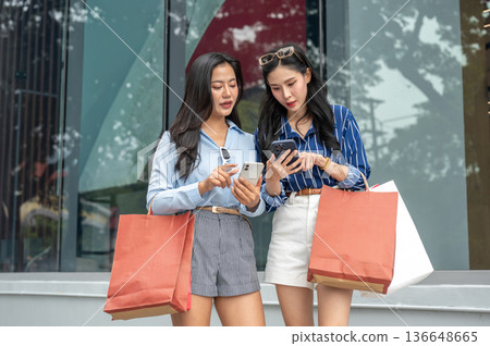 Pretty asian woman friend carrying shopping bag looking at their phone walking outside shopping mall Pretty asian woman friend carrying shopping bag looking at their phone walking outside shopping mall 136648665