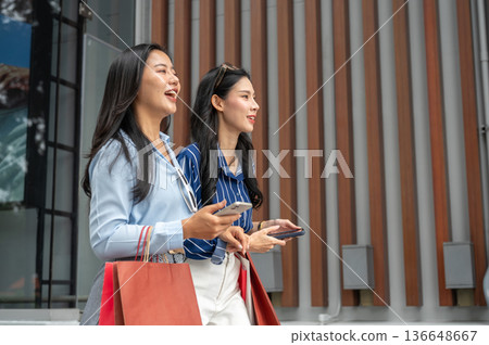 Asian woman friend holding phone carrying shopping bag while walking together outside shopping mall. 136648667