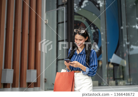 Pretty asian woman carrying shopping bags holding and looking at phone walking outside shopping mall 136648672