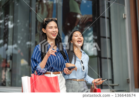 Pretty asian woman and laughing friend carrying shopping bag walking together outside shopping mall. 136648674