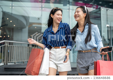 Pretty asian woman holding phone carrying shopping bag while walking with friend in front of a mall. 136648686