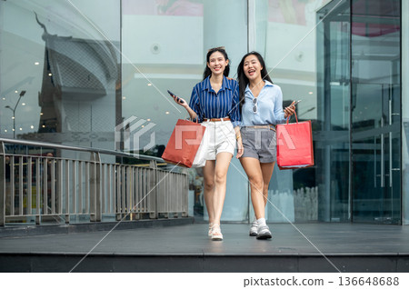 Pretty asian woman and friend holding phone carrying shopping bags while walking in front of a mall Pretty asian woman and friend holding phone carrying shopping bags while walking in front of a mall 136648688