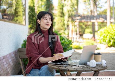 Asian woman holding pencil and book thinking aside laptop while sitting at wood table in garden cafe Asian woman holding pencil and book thinking aside laptop while sitting at wood table in garden cafe 136648838