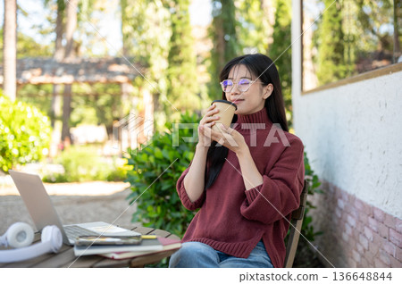 Glasses asian woman in sweater smelling coffee aroma while sitting at wooden table in garden cafe. Glasses asian woman in sweater smelling coffee aroma while sitting at wooden table in garden cafe. 136648844