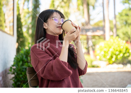 Glasses asian woman in sweater closing eyes smelling coffee aroma while sitting in the garden cafe. 136648845