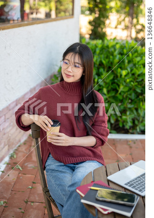 Glasses asian woman in sweater holding coffee aside laptop sitting at wooden table in garden cafe. Glasses asian woman in sweater holding coffee aside laptop sitting at wooden table in garden cafe. 136648846