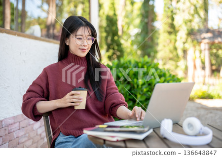 Glasses asian woman in sweater holding coffee looking at laptop sitting at wood table in garden cafe Glasses asian woman in sweater holding coffee looking at laptop sitting at wood table in garden cafe 136648847