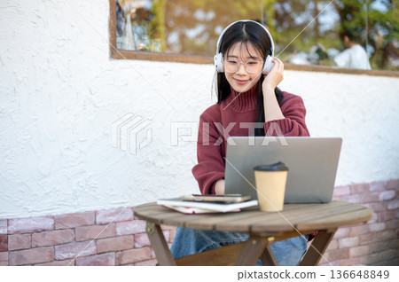 Glasses asian woman listening to music in headphones and looking at laptop on wooden table in cafe. Glasses asian woman listening to music in headphones and looking at laptop on wooden table in cafe. 136648849