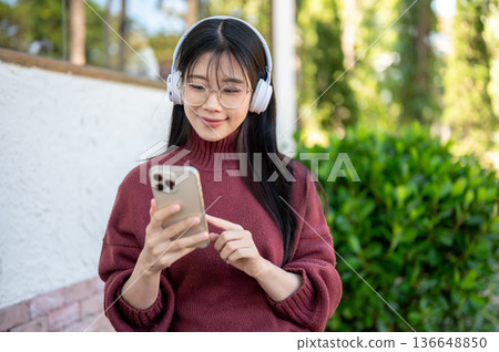 Glasses asian woman listening to music in headphones looking at phone while sitting in garden cafe. 136648850