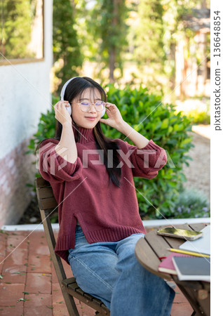 Glasses woman closing eyes listening to music in headphones sitting at wooden table in garden cafe. Glasses woman closing eyes listening to music in headphones sitting at wooden table in garden cafe. 136648854