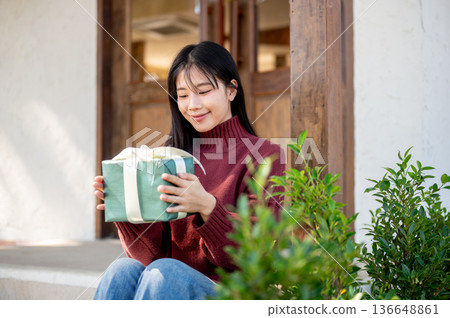 Asian woman in sweater looking at present gift box while sitting in front of cafe's door or entrance Asian woman in sweater looking at present gift box while sitting in front of cafe's door or entrance 136648861
