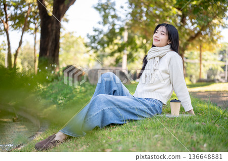 Asian woman in scarf leaning back sitting leisurely aside coffee on grass in garden or nature park. 136648881