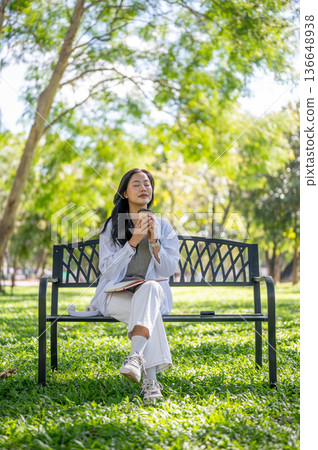 Asian woman closing eyes holding coffee cup while sitting on bench over green grass in garden park. 136648938