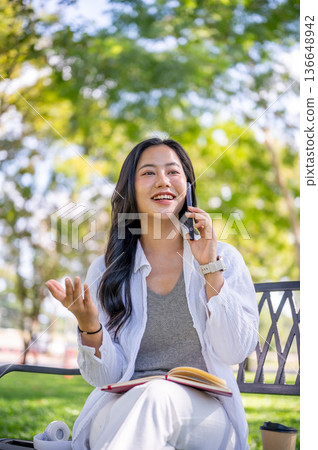 Asian woman holding and talking on smartphone over book while sitting on metal bench in garden park. Asian woman holding and talking on smartphone over book while sitting on metal bench in garden park. 136648942