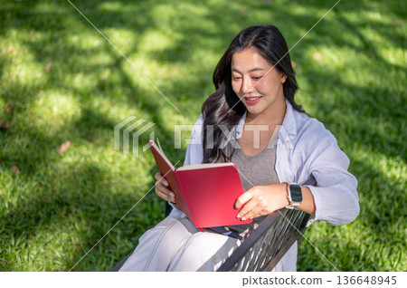 Asian woman holding and reading a book while sitting on metal bench over green grass in garden park. Asian woman holding and reading a book while sitting on metal bench over green grass in garden park. 136648945