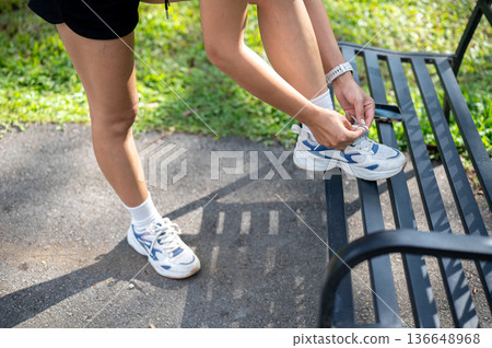 Close up of woman tying sneaker's shoelace on bench before exercise and standing on pavement in park Close up of woman tying sneaker's shoelace on bench before exercise and standing on pavement in park 136648968