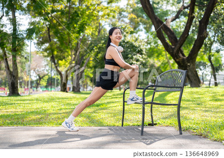 Asian woman in sportswear stretching leg on bench before exercise while standing on pavement in park 136648969