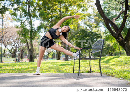 Asian woman in sportswear standing stretching muscles before exercise on bench over pavement in park 136648970