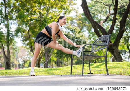 Asian woman in sportswear standing stretching muscle before exercise on bench over pavement in park. 136648971