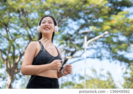 Asian woman in sportswear listening to music in headphones and exercise by running jogging in park. Asian woman in sportswear listening to music in headphones and exercise by running jogging in park. 136648982