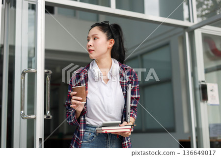 Asian woman student holding coffee with calculator on books walking out of a building's glass door. Asian woman student holding coffee with calculator on books walking out of a building's glass door. 136649017