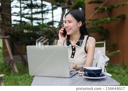 Asian woman student talking on phone aside laptop while sitting at table on grass in outdoors cafe. 136649027