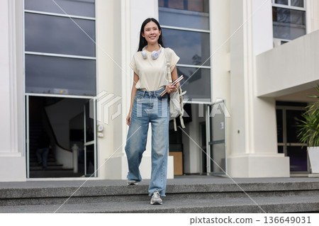 Asian woman student holding book and carry a bag while walking down stairs in front of a building 136649031