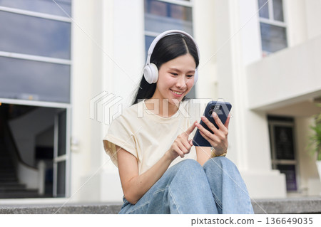 Asian woman student looking at phone listening to music as sitting in front of a library or building Asian woman student looking at phone listening to music as sitting in front of a library or building 136649035
