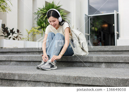 Asian woman student listening to music in headphone tying shoelace as sitting in front of a building Asian woman student listening to music in headphone tying shoelace as sitting in front of a building 136649038