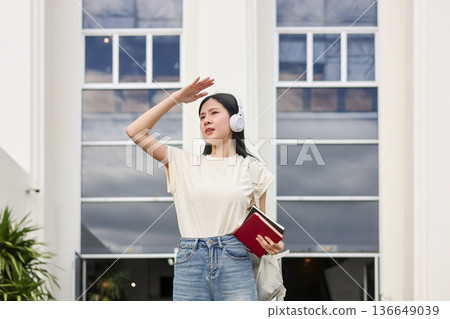 Asian woman student holding book using hand covering eye from sunlight standing in front of building Asian woman student holding book using hand covering eye from sunlight standing in front of building 136649039