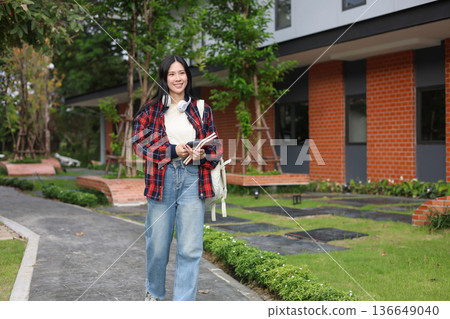 Asian woman college student holding phone and books while walking on sidewalk beside garden or park. 136649040