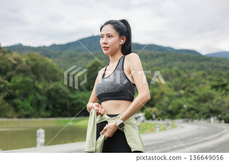 Asian woman in sportswear tying jacket on waist before exercise and standing on street road in park. 136649056