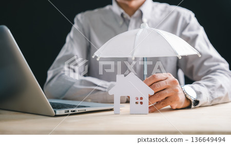 Human hand provides important home insurance protection concept for house, holding white umbrella safeguarding property model, symbolizing security and financial care on desk near laptop Human hand provides important home insurance protection concept for house, holding white umbrella safeguarding property model, symbolizing security and financial care on desk near laptop 136649494