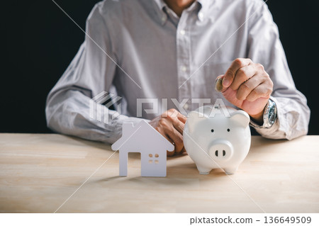 Estate property investment concept, person carefully saving money for future home ownership, placing coin in white piggy bank, showing financial planning for real house dream 136649509