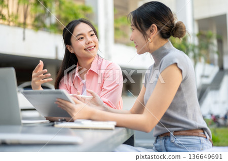 Asian woman student talking to friend holding tablet working on group project at table in university Asian woman student talking to friend holding tablet working on group project at table in university 136649691