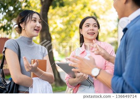 Asian woman student holding book talking to man and friend while standing beside university's garden Asian woman student holding book talking to man and friend while standing beside university's garden 136649714