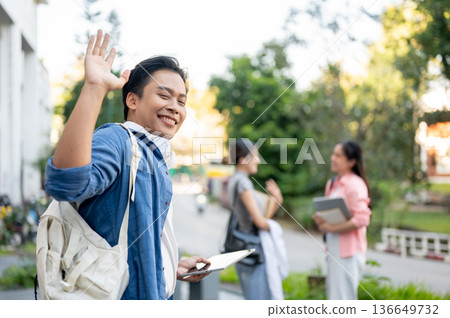 Happy asian man student holding tablet waving a hand while walking to woman friends in university Happy asian man student holding tablet waving a hand while walking to woman friends in university 136649732