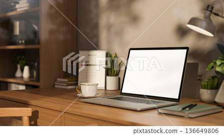 White screen laptop and coffee with lamp and calendar on wooden table aside bookshelf in study room. White screen laptop and coffee with lamp and calendar on wooden table aside bookshelf in study room. 136649806