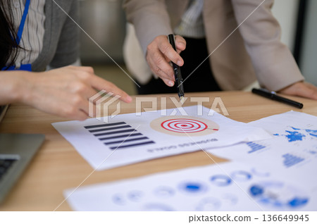 Close up of businesswoman's hand pointing pen at document on wooden table aside coworker in office. 136649945