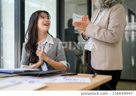 Old asian businesswoman clapping hand and talking with young coworker at wooden table in office room Old asian businesswoman clapping hand and talking with young coworker at wooden table in office room 136649947