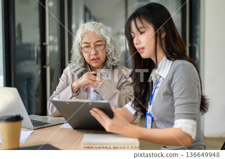 Old asian businesswoman looking at clipboard and talking to young coworker at wooden table in office Old asian businesswoman looking at clipboard and talking to young coworker at wooden table in office 136649948