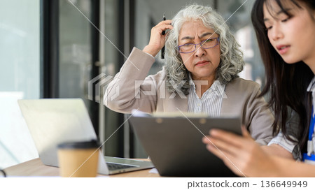 Old asian businesswoman looking confused at clipboard aside young coworker at wooden table in office Old asian businesswoman looking confused at clipboard aside young coworker at wooden table in office 136649949