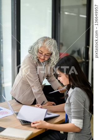 Old asian businesswoman sitting on table looking at laptop as young coworker writing paper in office 136649957