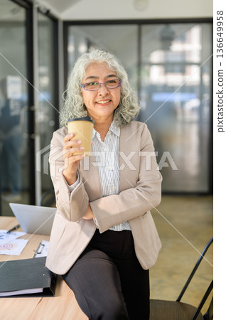 Old asian businesswoman holding coffee cup while standing or sitting leaning on table in office. 136649958