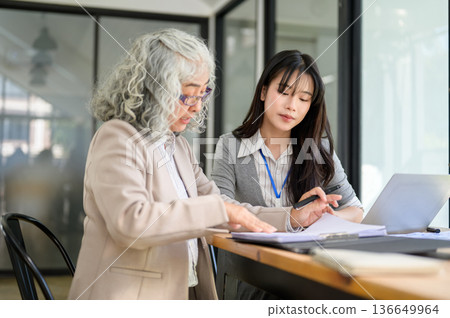 Old asian businesswoman holding pen explaining document to young coworker while sitting in office. Old asian businesswoman holding pen explaining document to young coworker while sitting in office. 136649964