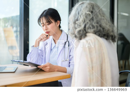 Asian woman doctor holding pen looking at clipboard sitting with old patient at table in diagnosis room 136649969